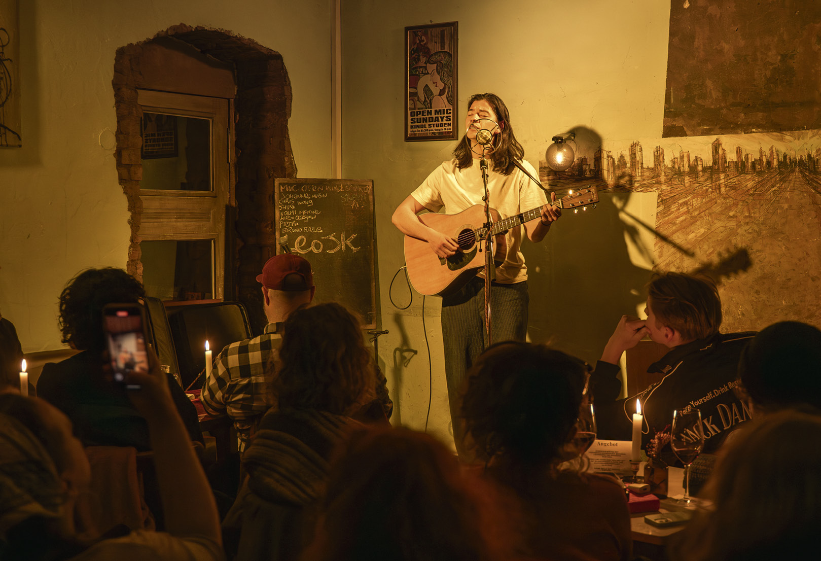 Eine Person singt und macht Musik mit einer Guitarre auf einer Bühne in einer kleinen Bar.