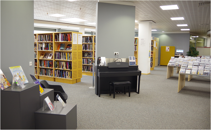 Eine Bibliothek mit vielen Gelben Bücherregalen, ein Schwarzes Klavier und Austellungsblöcke mit Filmen und Büchern.