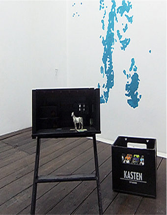 Eine Kunstausstellung in einer Ecke, weiße Wände mit einem Spritzer blauer Farbe, rechts gibt es ein schwarzer Glasskasten und ein schwarzer Tritthocker worauf eine schwarze box steht und innen ein kleines weißes Pferd steht.